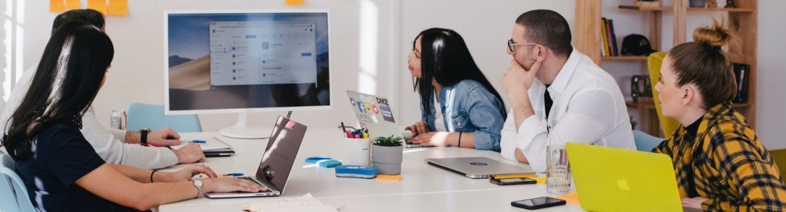 Fünf Personen sitzen an einem Tisch und schauen auf einen Monitor.